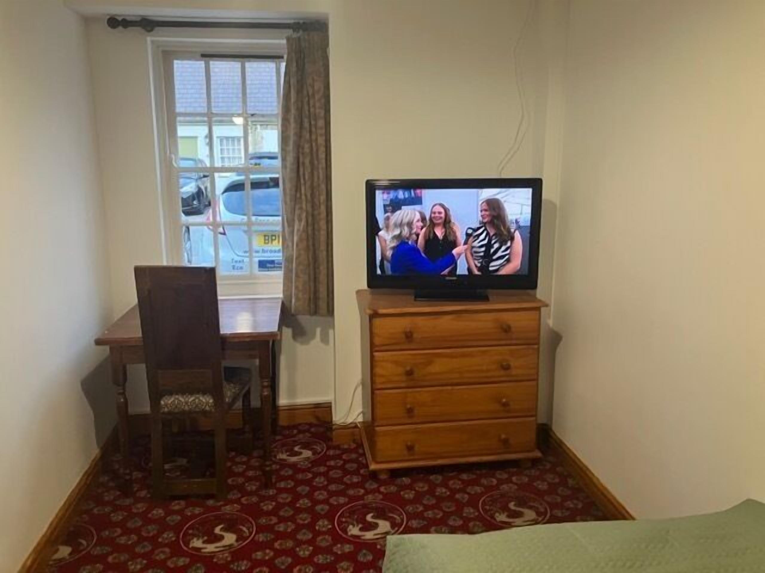Een kamer met een televisie op een houten dressoir, een stoel, een raam met uitzicht op geparkeerde auto's en een kamerbreed tapijt met dessin.