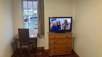 Een kamer met een televisie op een houten dressoir, een stoel, een raam met uitzicht op geparkeerde auto's en een kamerbreed tapijt met dessin.