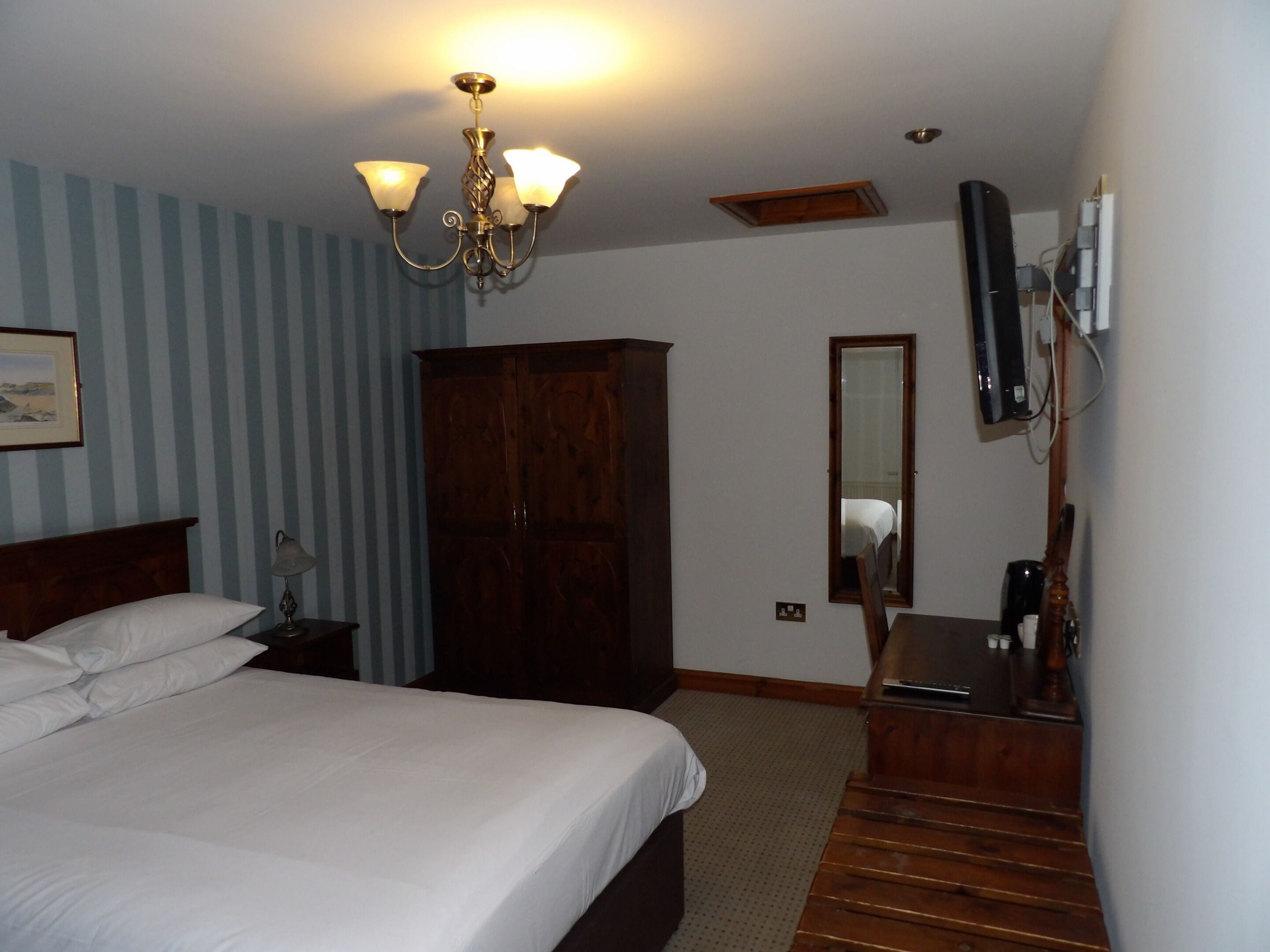 A hotel room with a bed, a wooden wardrobe, a desk with a chair, a mirror, and a television mounted on the wall.