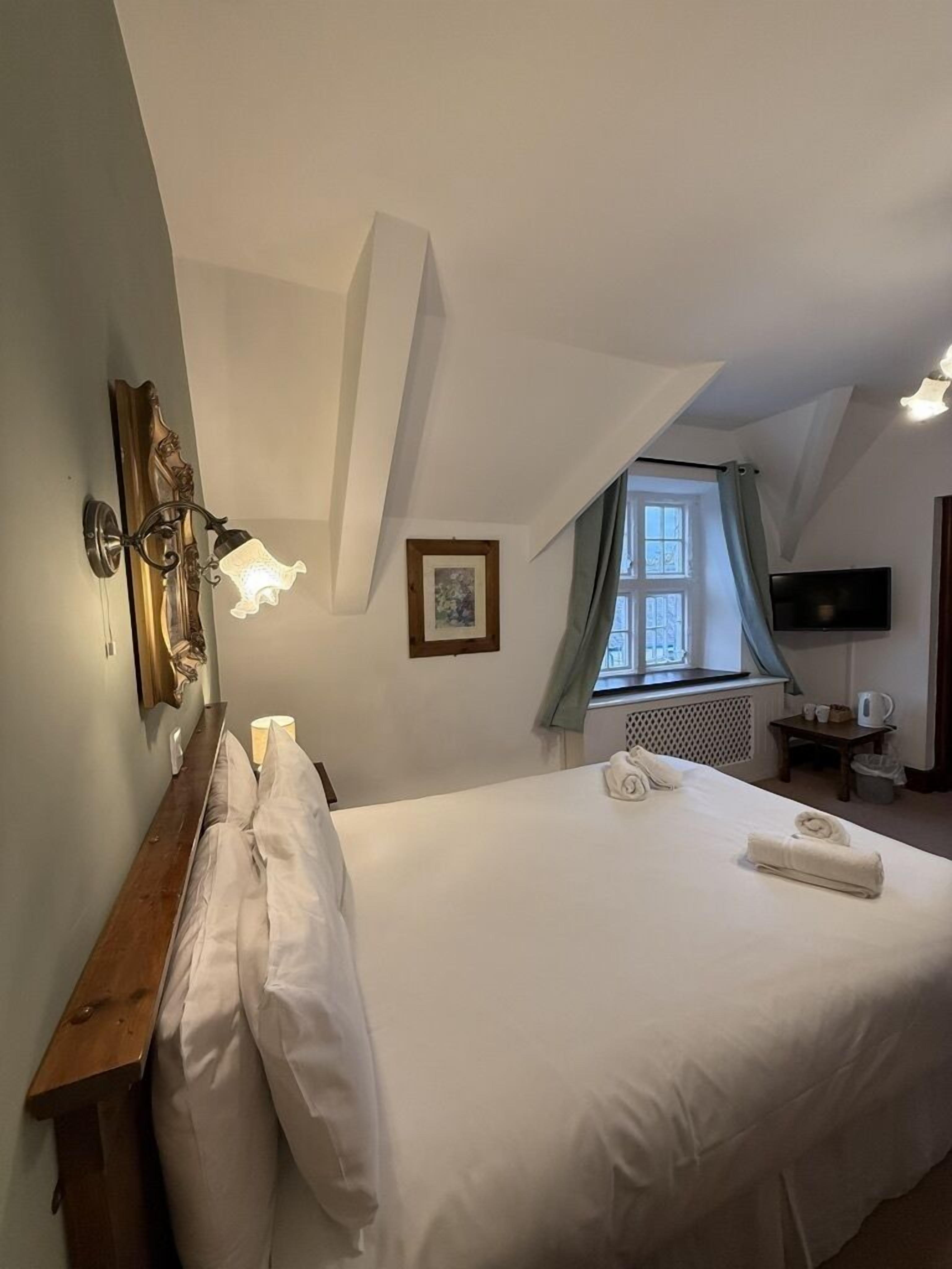 Un dormitorio con una cama grande, una mesita de noche de madera, una lámpara de pared, una ventana con cortinas y un televisor en la pared.