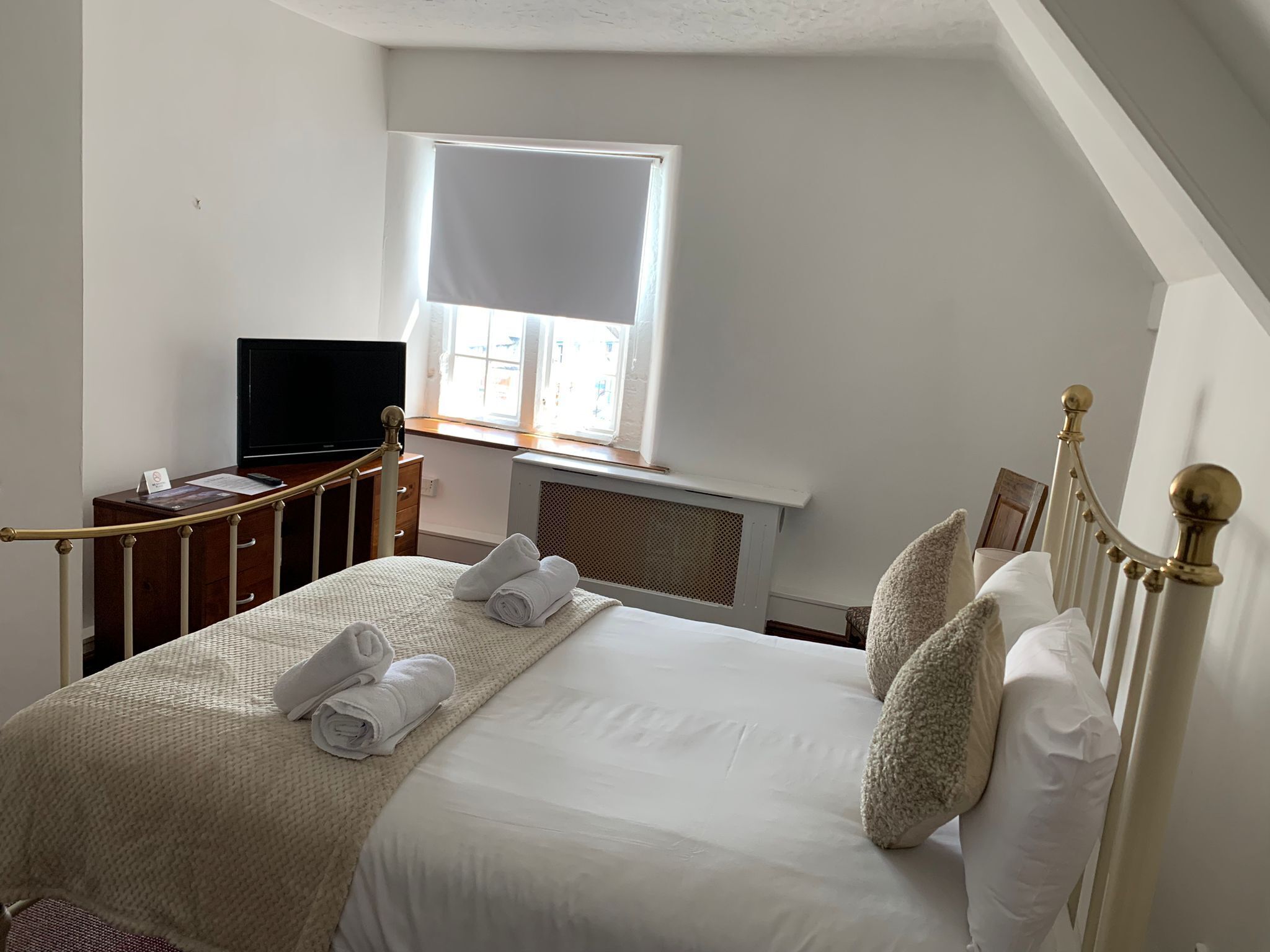 A neatly made bed with white linens and rolled towels, a television on a wooden dresser, and a window with a white blind.
