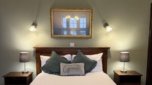 Cama bem arrumada com colcha verde e duas almofadas, uma com mensagem para hóspedes. Há duas luminárias de cabeceira e um quadro emoldurado acima da cama.