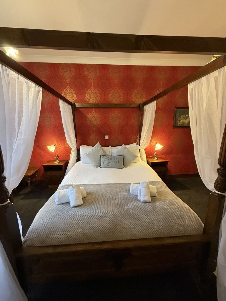 A four-poster canopy bed with white linens, two bedside lamps, and a red patterned wall.