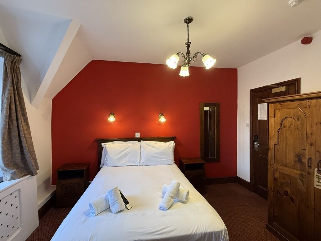 A hotel room with a bed, two bedside tables, a wooden wardrobe, a red accent wall, and a chandelier.