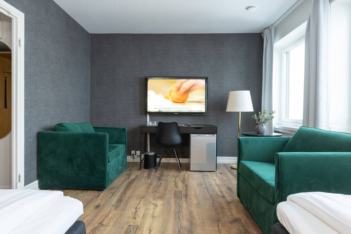A modern hotel room with a grey wall, wooden flooring, a desk with a computer, a green sofa, and a TV mounted on the wall.