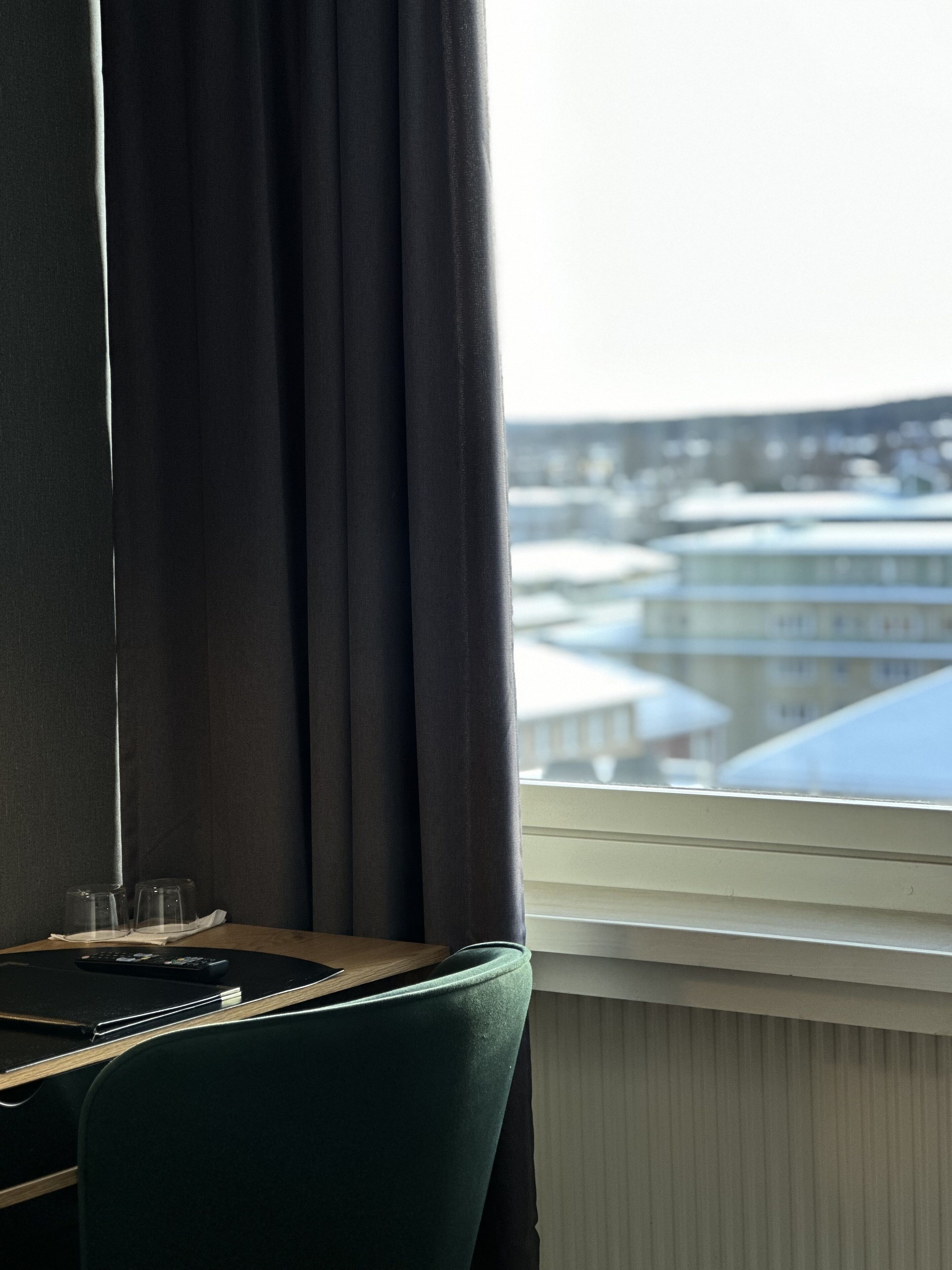 A hotel room with a view of buildings through a window, a desk with a chair, and a window blind.
