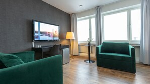 Una habitación de hotel moderna con televisión de pantalla plana, un escritorio con silla, un sofá, una mesita con lámpara y un ventanal con cortinas.