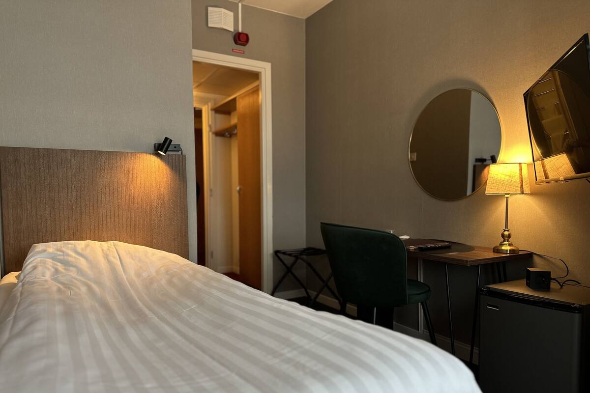A hotel room with a bed, a desk, a chair, a desk lamp, a small TV, and a mirror on the wall.