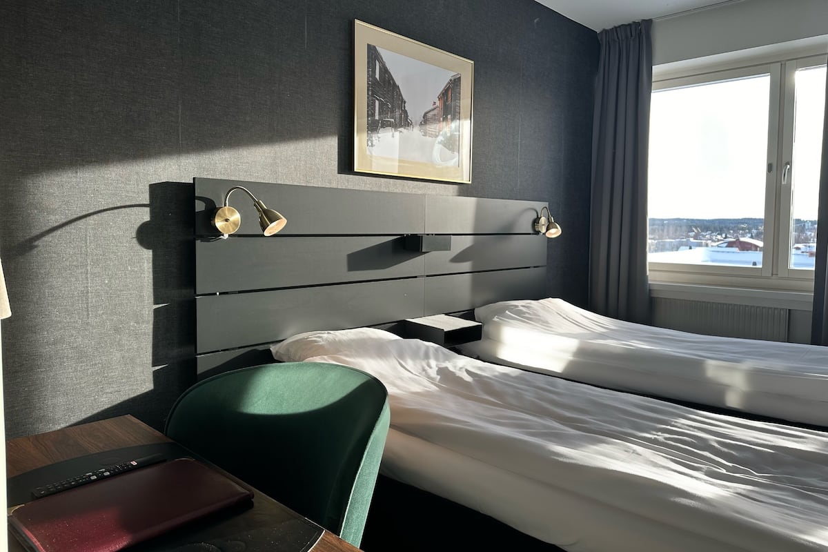 A hotel room with two beds, a desk with a laptop, a chair, and a window with a view of a snowy landscape.