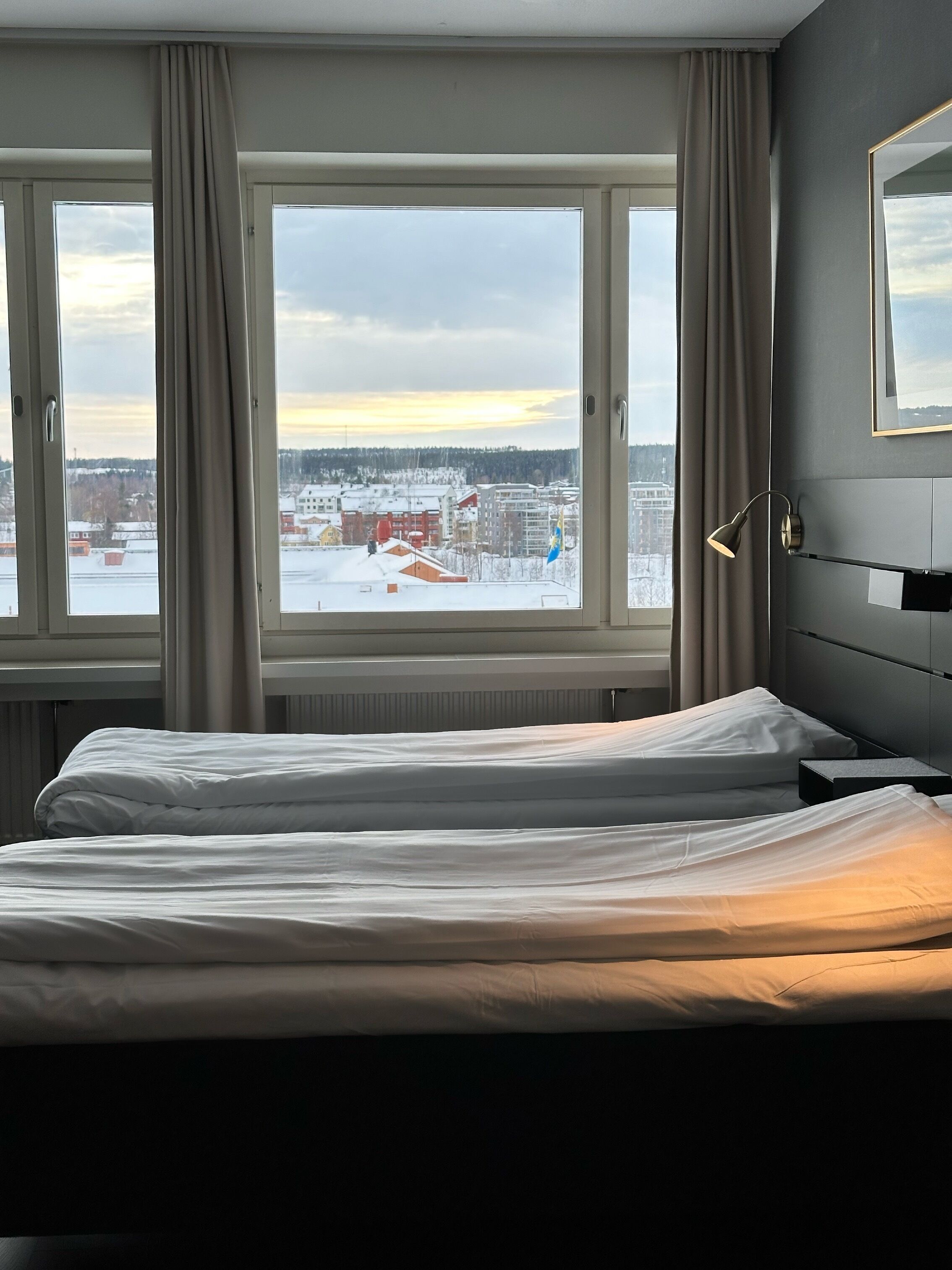 A hotel room with two beds, a large window with curtains, a bedside lamp, and a view of a snowy cityscape.