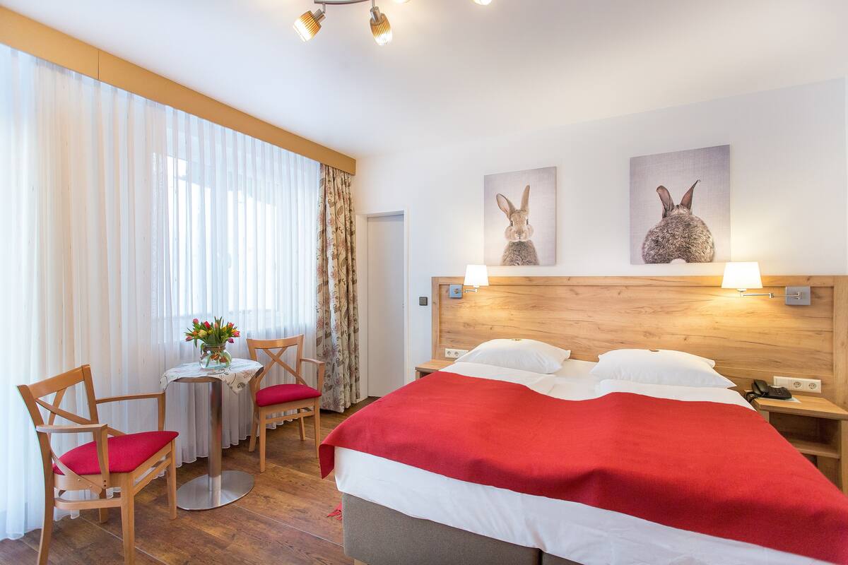 A hotel room with a large bed, a wooden headboard, two wall-mounted lamps, a small table with a chair, and two framed pictures of rabbits.