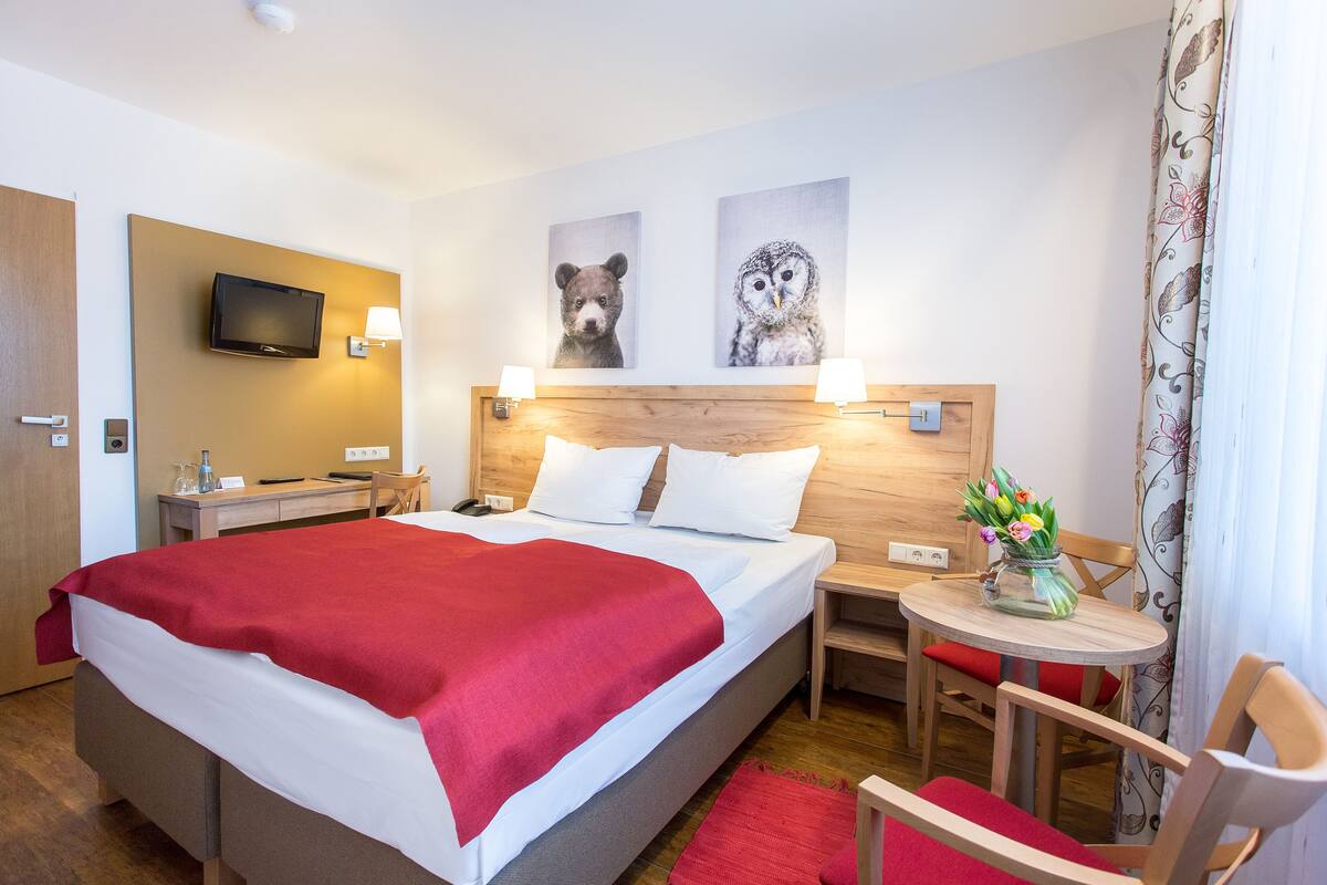 A hotel room with a bed, bedside tables, a TV, artwork, a small table with a vase of flowers, and a chair.