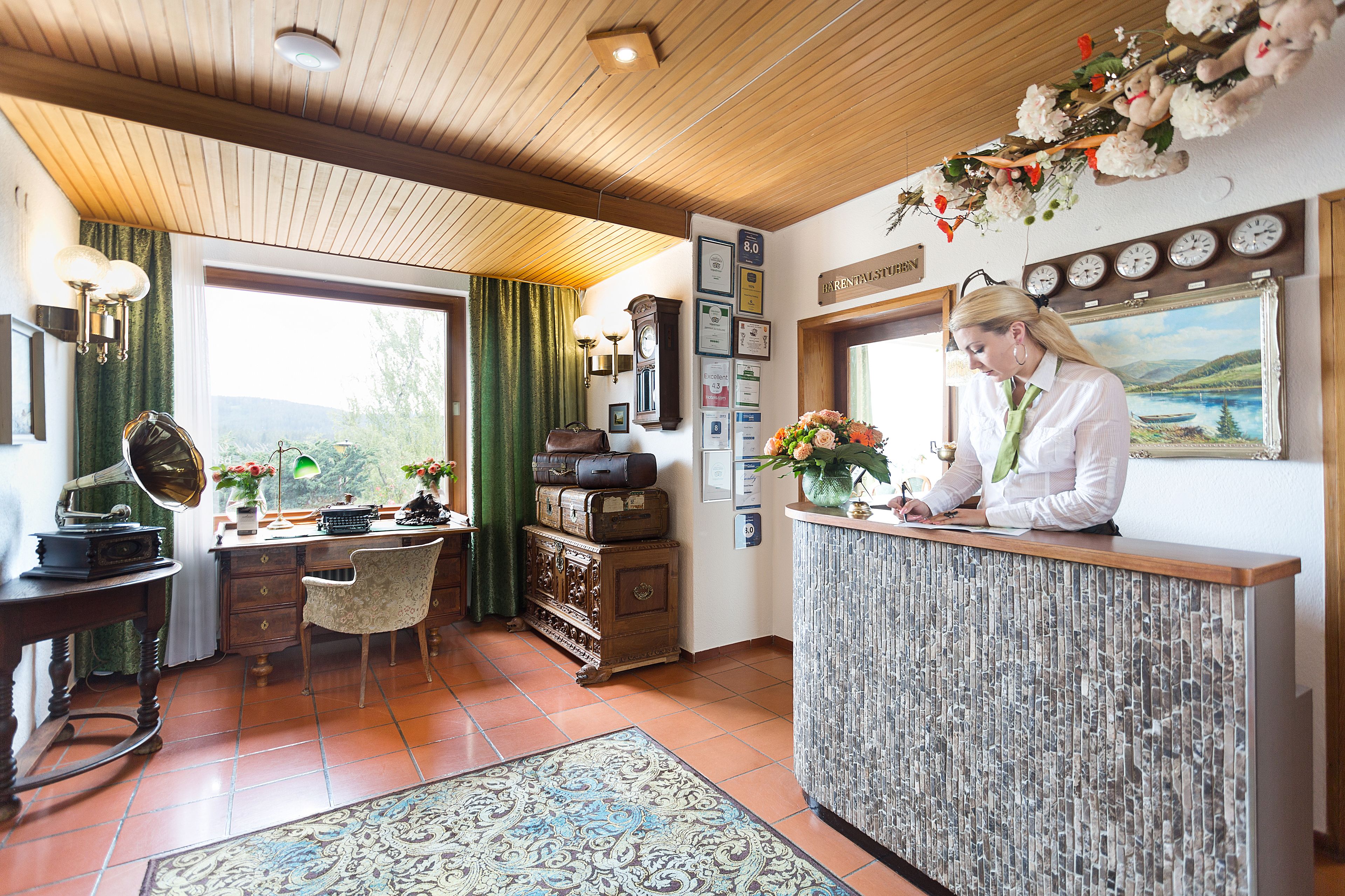 A reception desk with a receptionist, a vintage radio, a desk with a typewriter, a chair, a cabinet with suitcases, and a view of the outdoors.