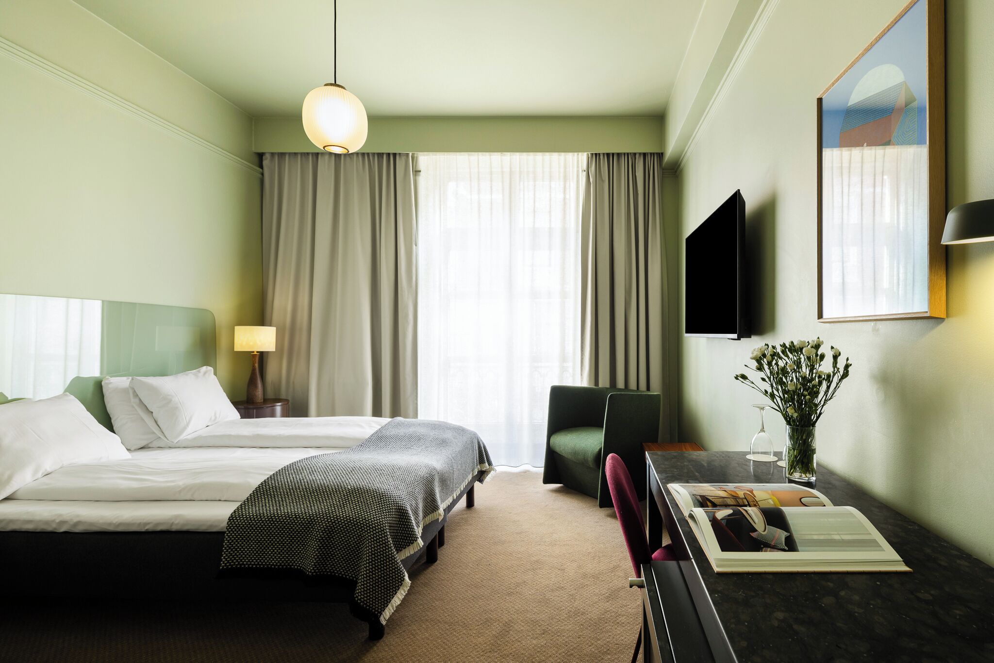 Een hotelkamer met een bed, een bureau met stoel, een televisie, een lamp en een raam met gordijnen.
