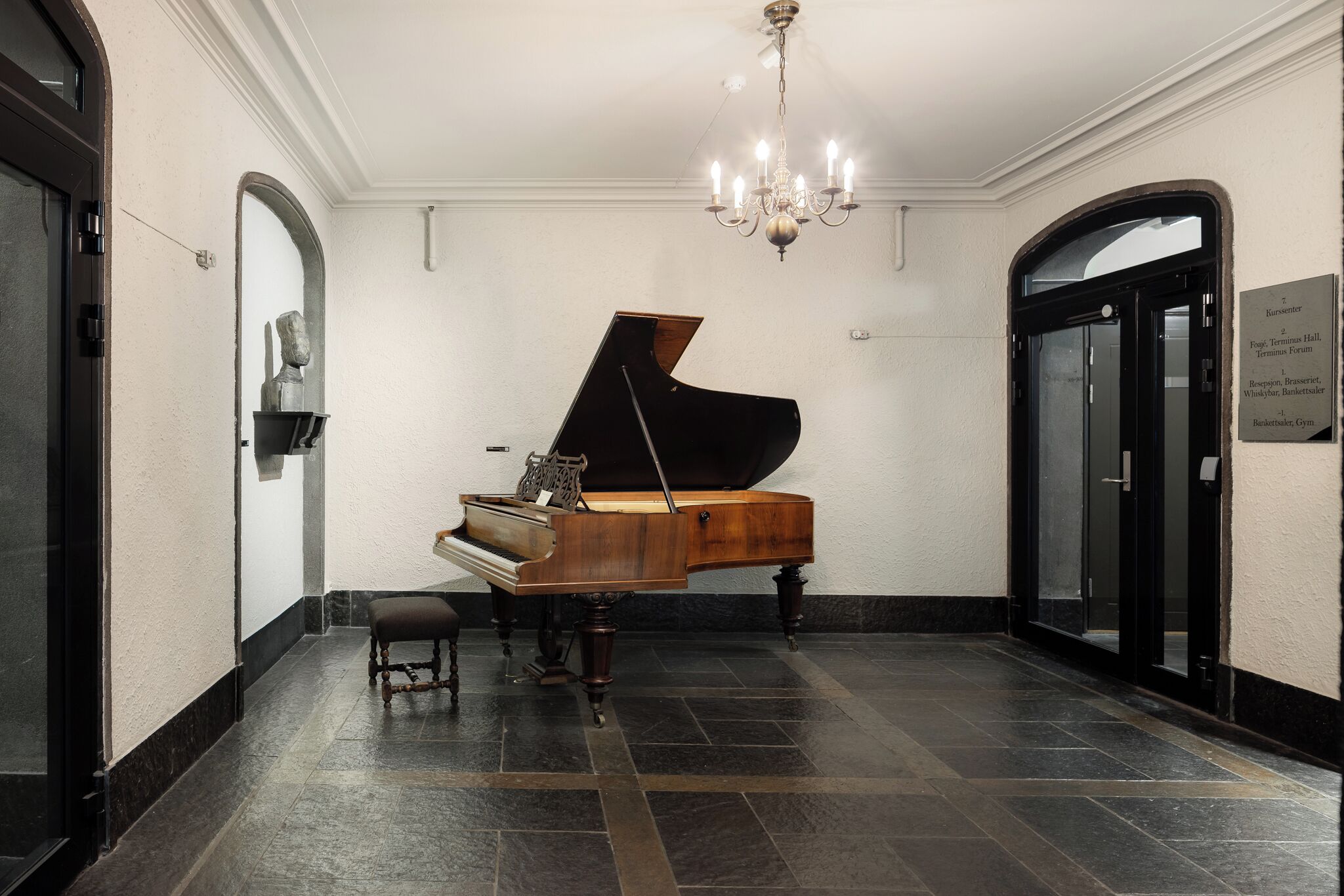 A grand piano in a spacious hallway with a chandelier, a bench, and a sign indicating the location.