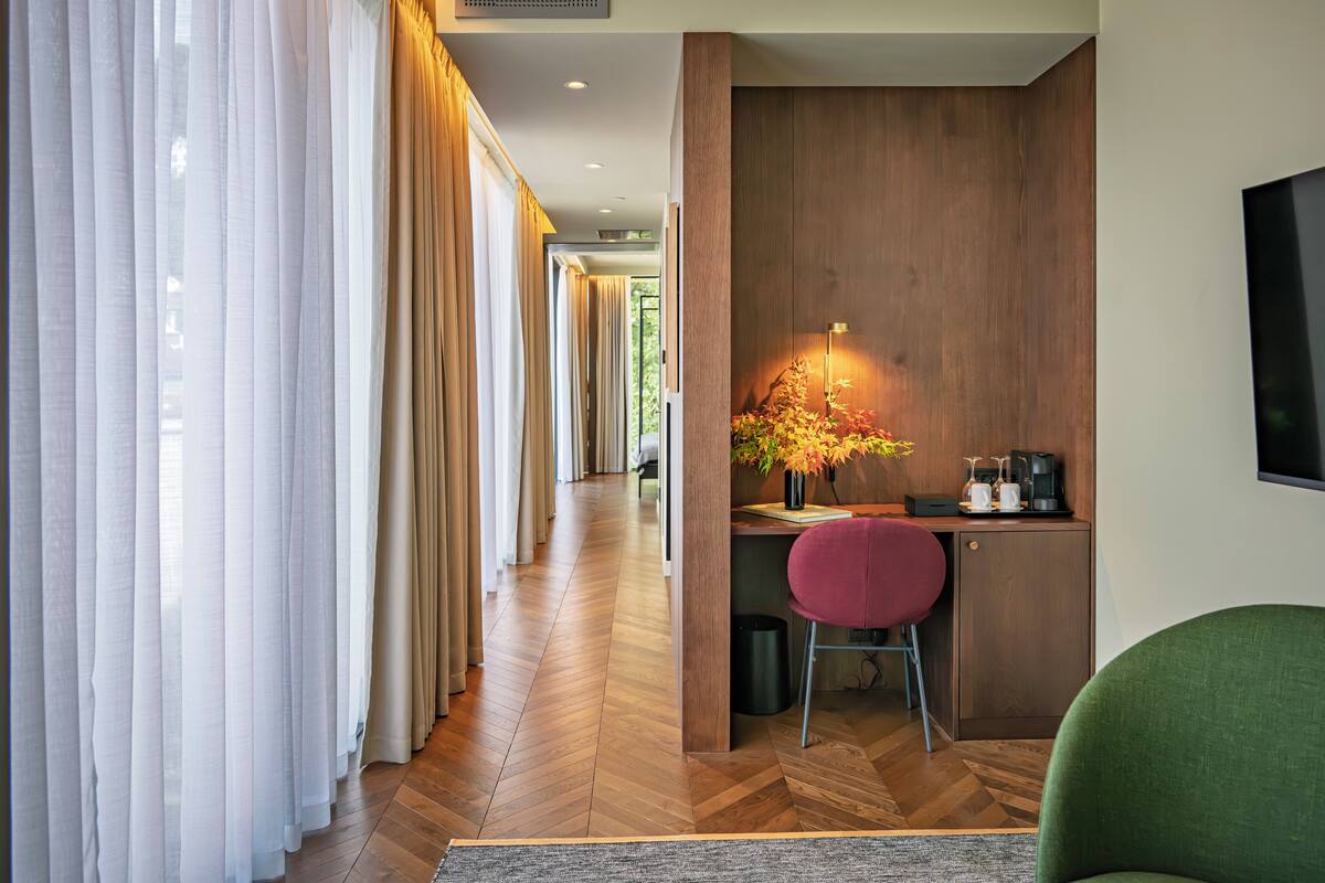Ein modernes Hotelzimmer mit Holzboden im Fischgrätmuster, einem Schreibtisch mit Stuhl, einer kleinen Küchenzeile und einem großen Fenster mit durchsichtigen Vorhängen.