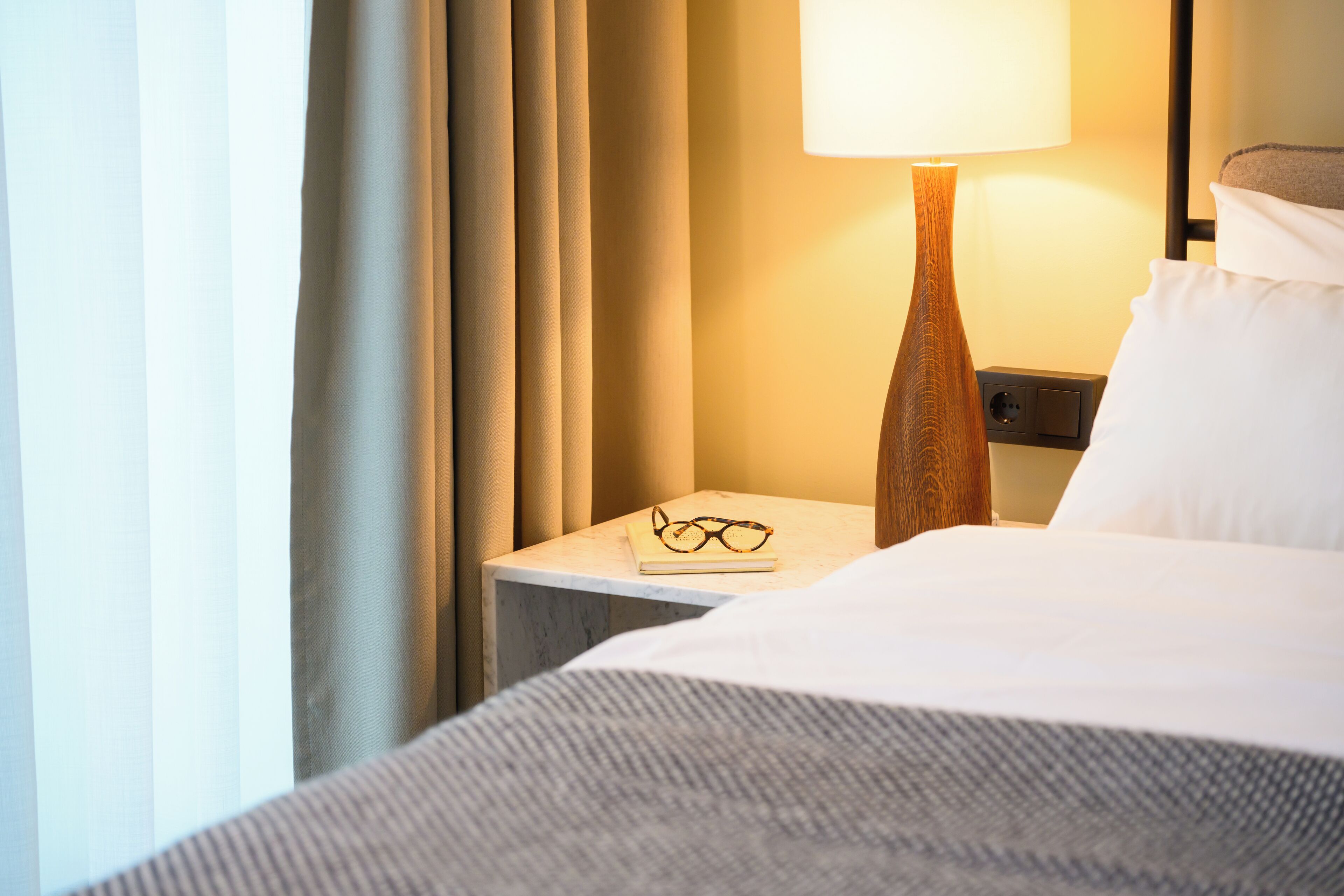Una cama bien tendida con un almohadón blanco, una mesita de noche de madera con una lámpara y un par de gafas sobre la mesa.