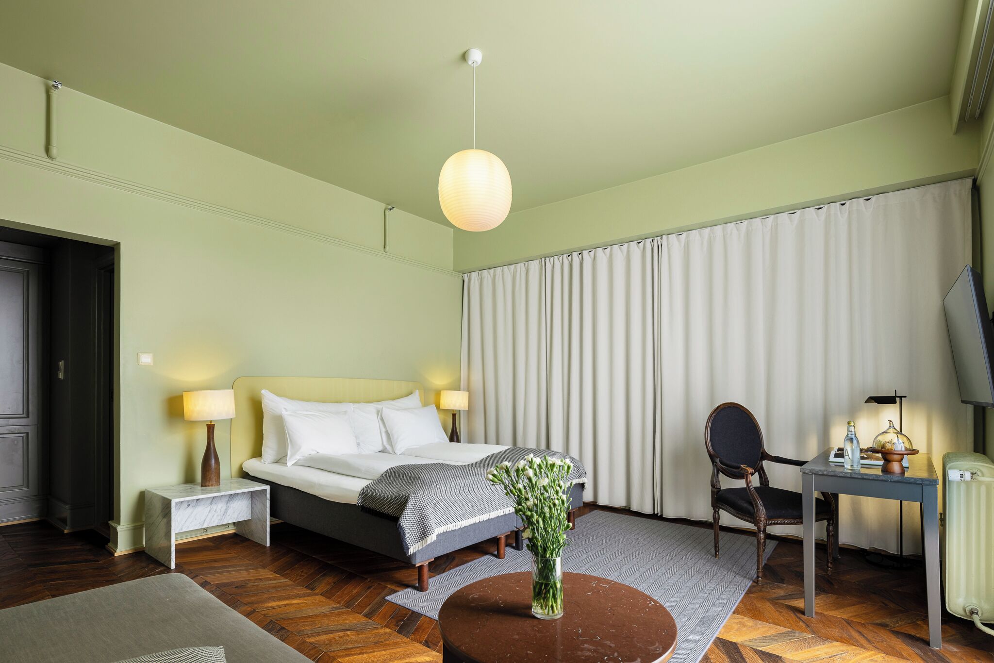 Habitación de hotel con una cama, lámparas de noche, un escritorio con una silla, un jarrón con flores y un radiador.