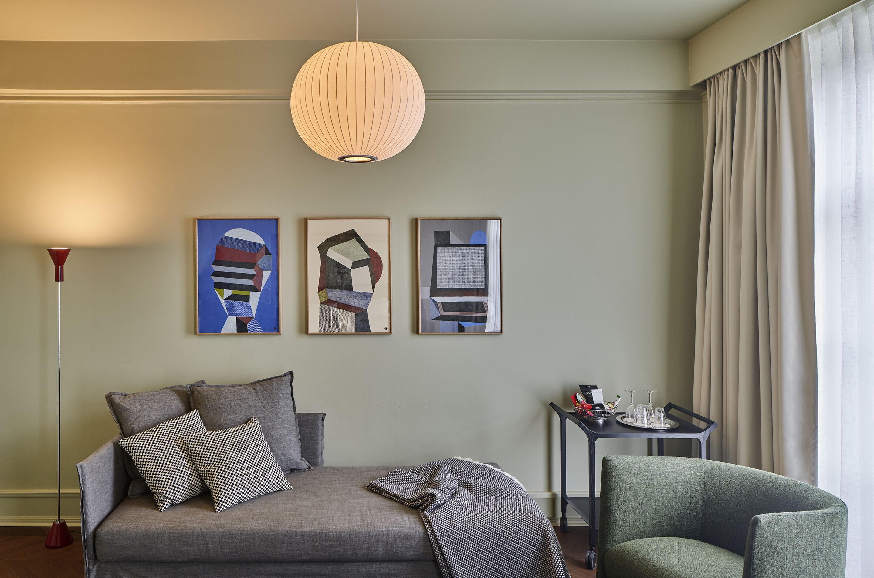 A modern hotel room with a grey sofa, a green armchair, and a small table with a tray of glasses and a red object.