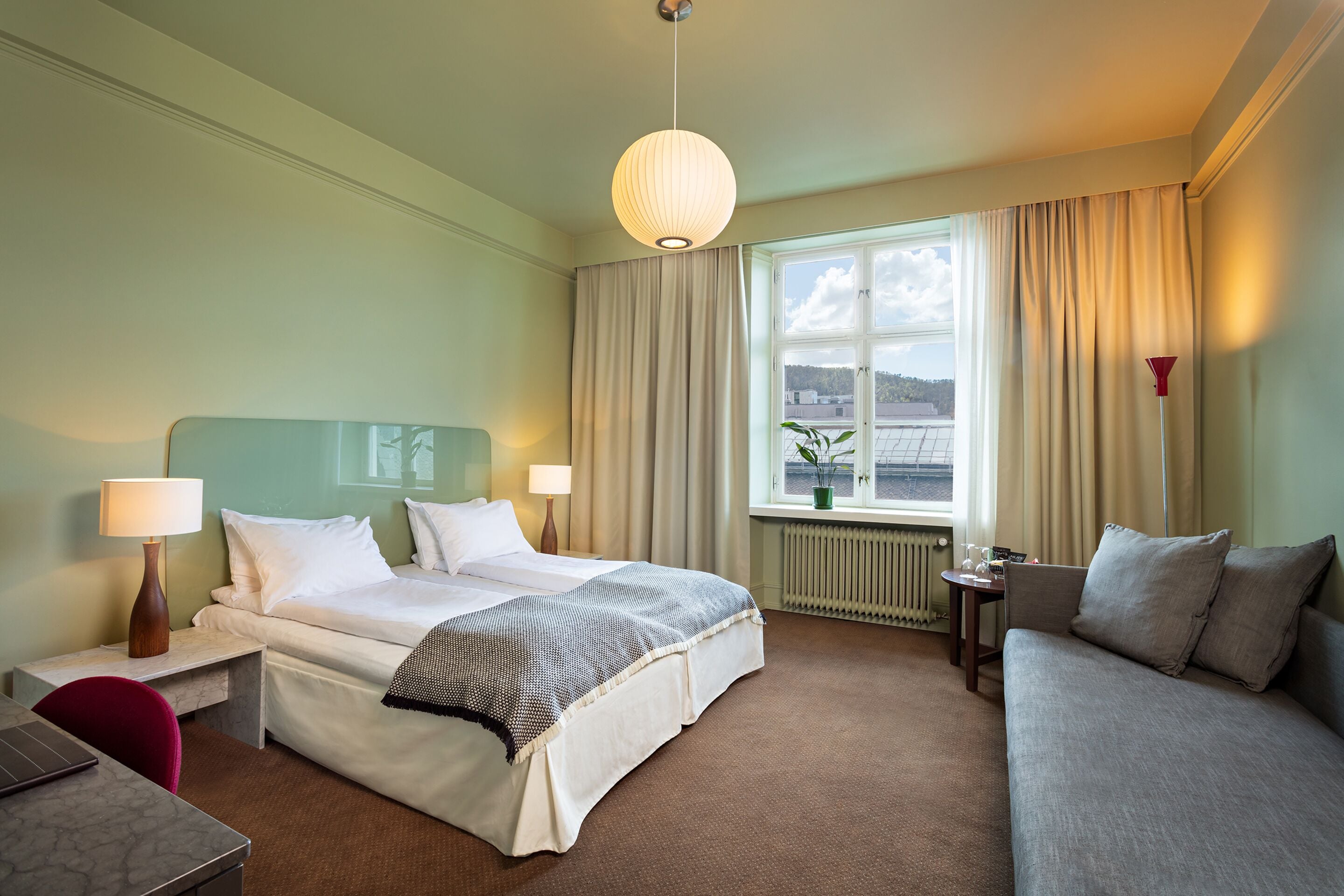 A hotel room with a bed, a sofa, a desk with a lamp, a window with curtains, and a view of the outside.