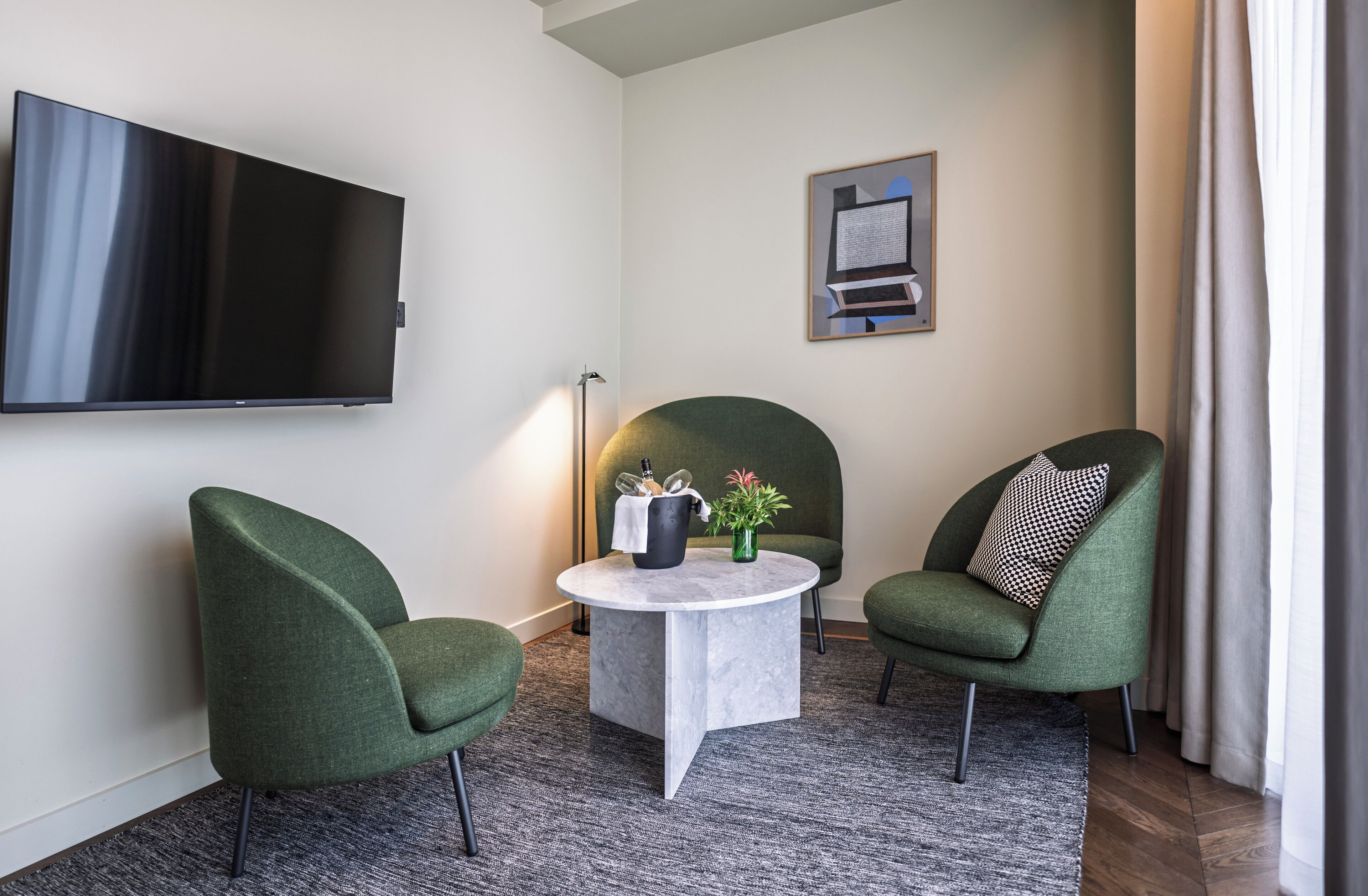 Una sala de estar moderna con un televisor de pantalla plana, dos sillones verdes, una mesa de centro redonda con un jarrón y una taza negra, y un cuadro enmarcado en la pared.