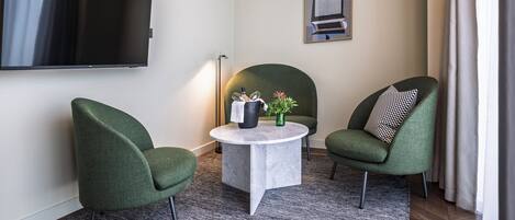 Una sala de estar moderna con un televisor de pantalla plana, dos sillones verdes, una mesa de centro redonda con un jarrón y una taza negra, y un cuadro enmarcado en la pared.