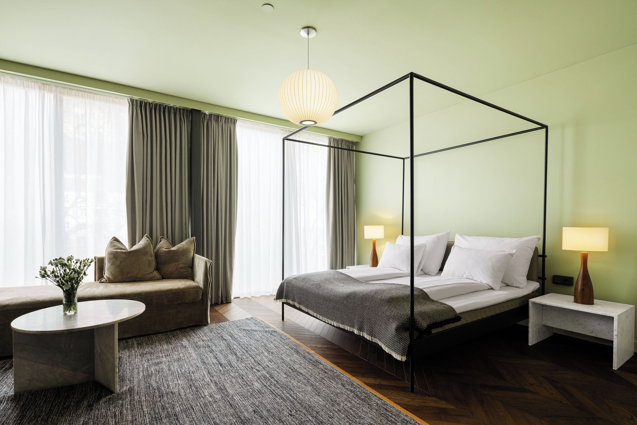 A modern bedroom with a four-poster bed, a sofa, a round coffee table, and a nightstand with a lamp.