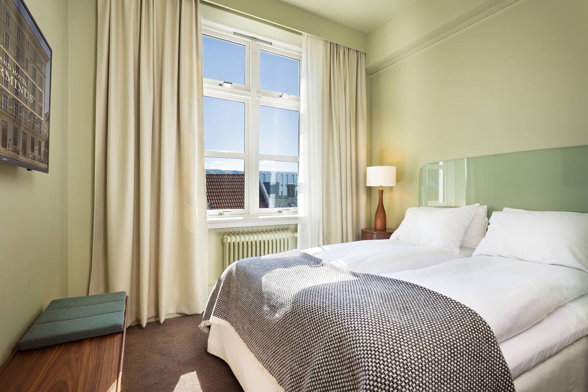 Ein Hotelzimmer mit einem großen Bett, einem Fenster mit Vorhängen, einem Heizkörper, einem Nachttisch mit Lampe und einem Wandbild.