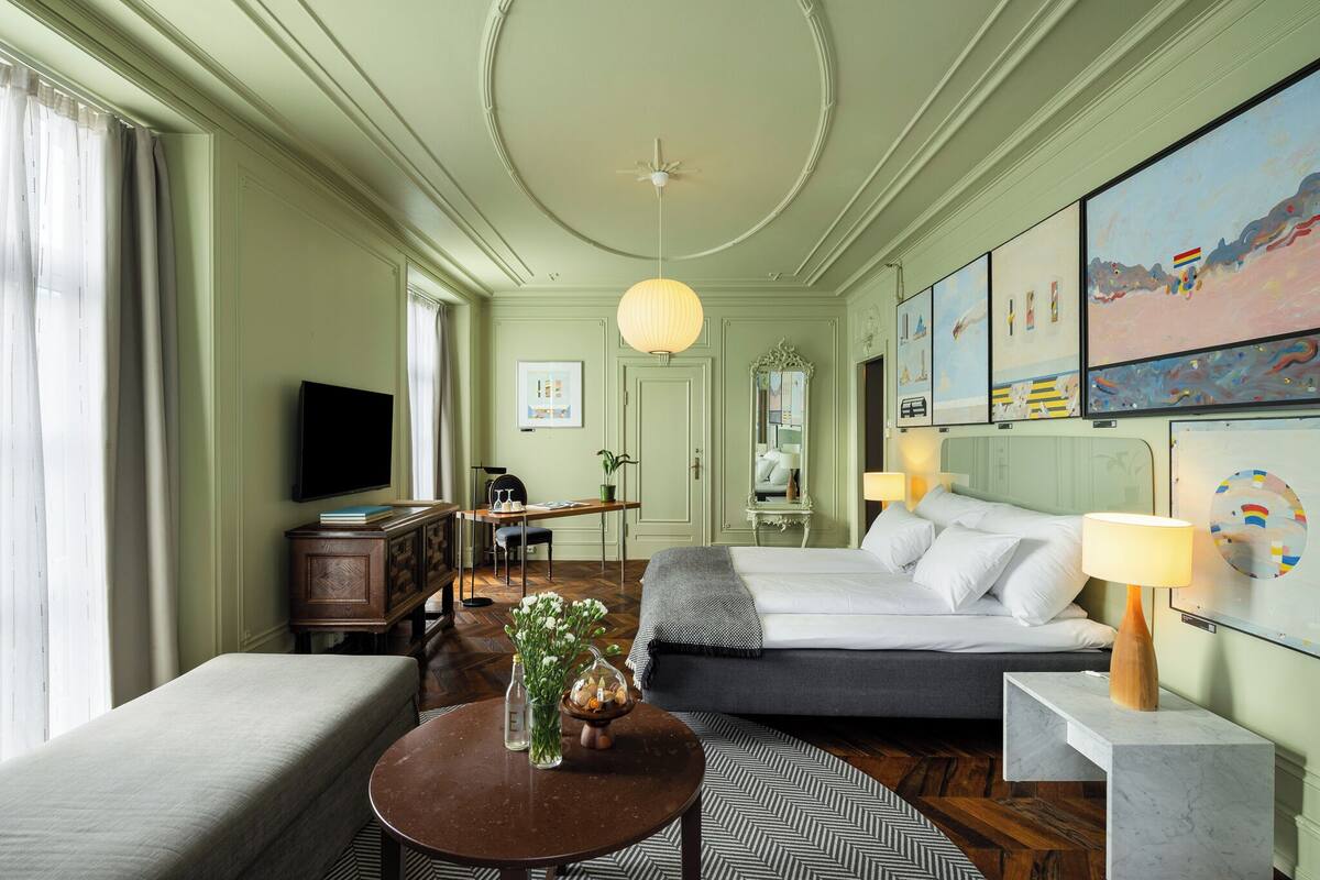 Ein Hotelzimmer mit Bett, Nachttisch, Lampe, Fernseher, Schreibtisch, Stuhl und Kunstwerken an den Wänden.