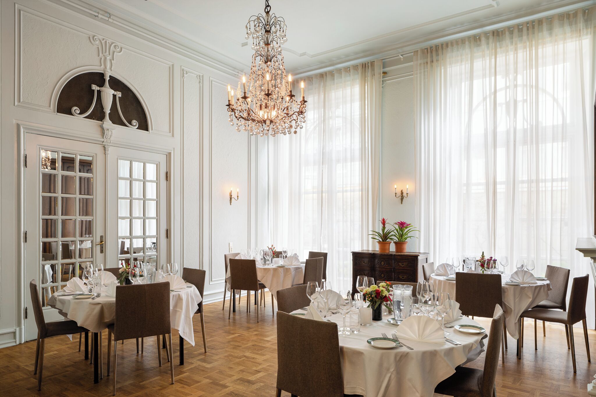 A formal dining room with chandeliers, white curtains, and a wooden floor.