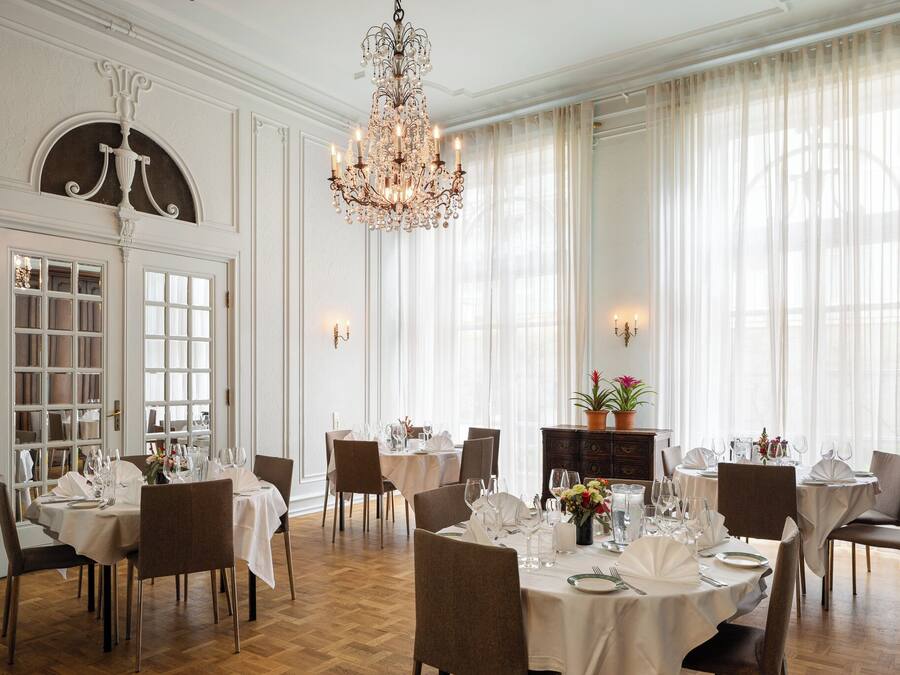 A formal dining room with chandeliers, white curtains, and a wooden floor.