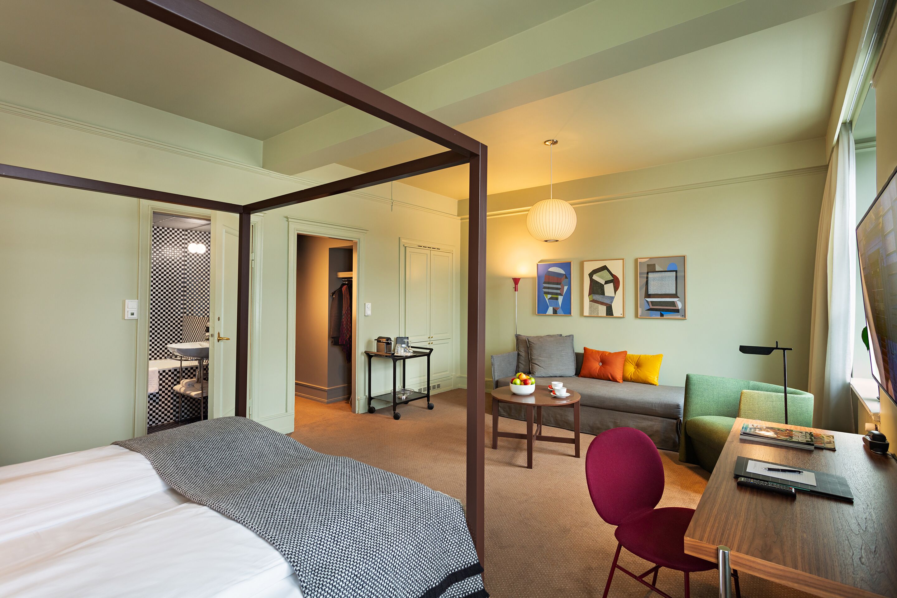 Hotelkamer met een bed, een bureau, een stoel, een bank en een eettafel. Er is een raam met gordijnen, een aan de muur bevestigde televisie en een deur naar een andere kamer.