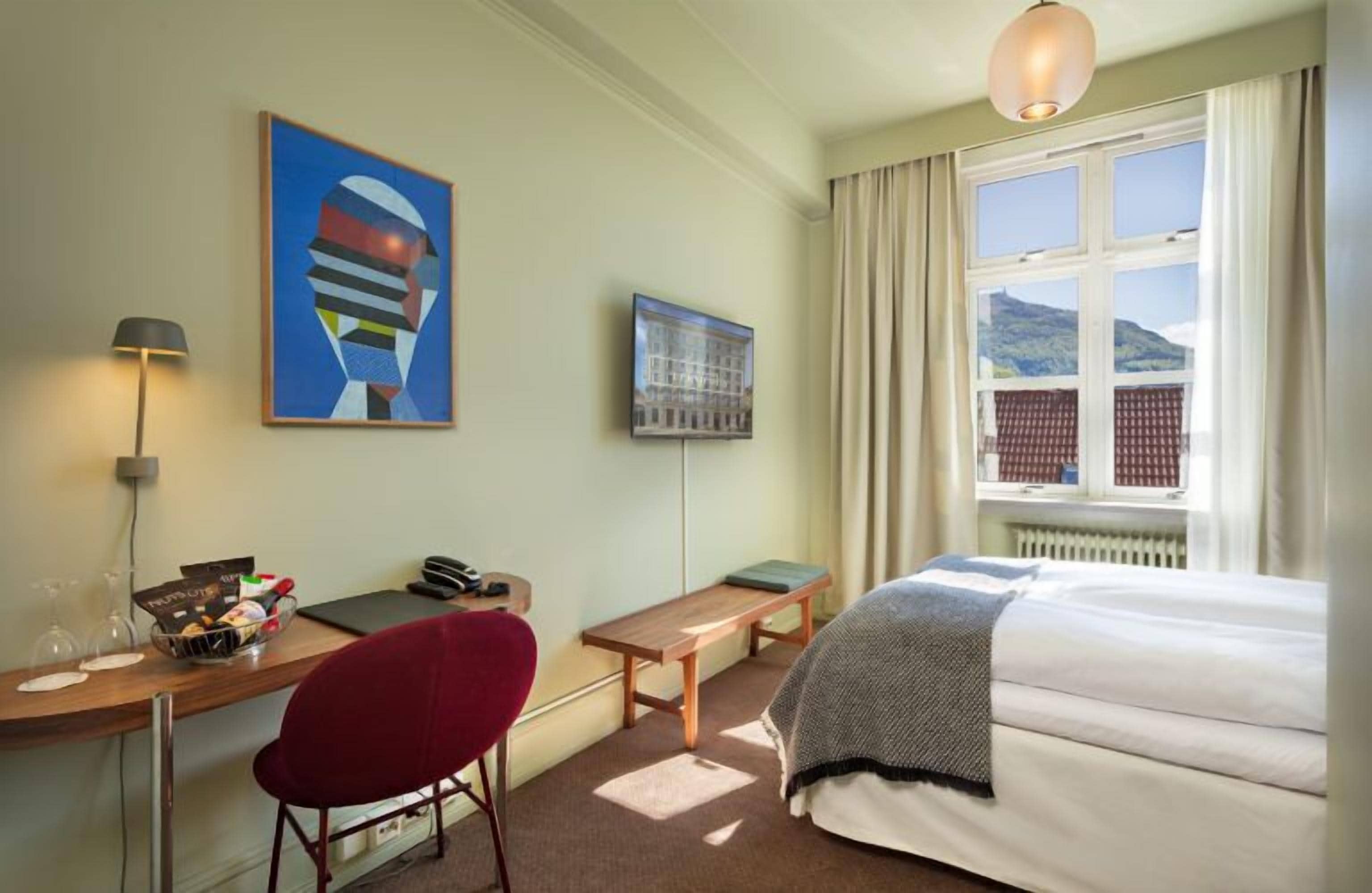 A hotel room with a bed, a desk with a lamp, a chair, a window with curtains, and artwork on the walls.