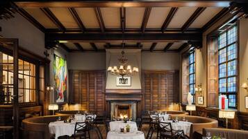 A dining room with round tables, wooden paneling, a fireplace, and large windows.