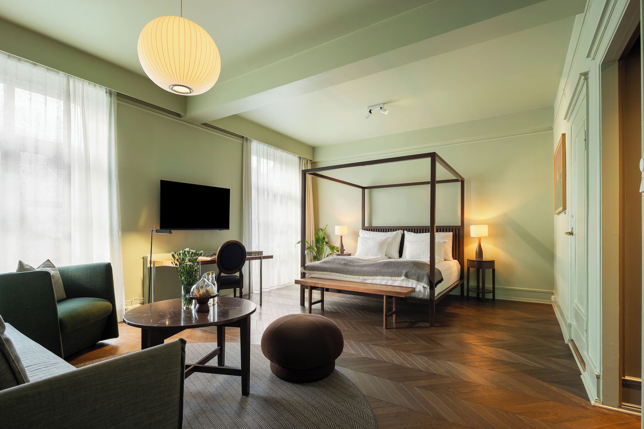 A modern hotel room with a four-poster bed, a flat-screen TV, a sofa, a dining table, and a large window with sheer curtains.
