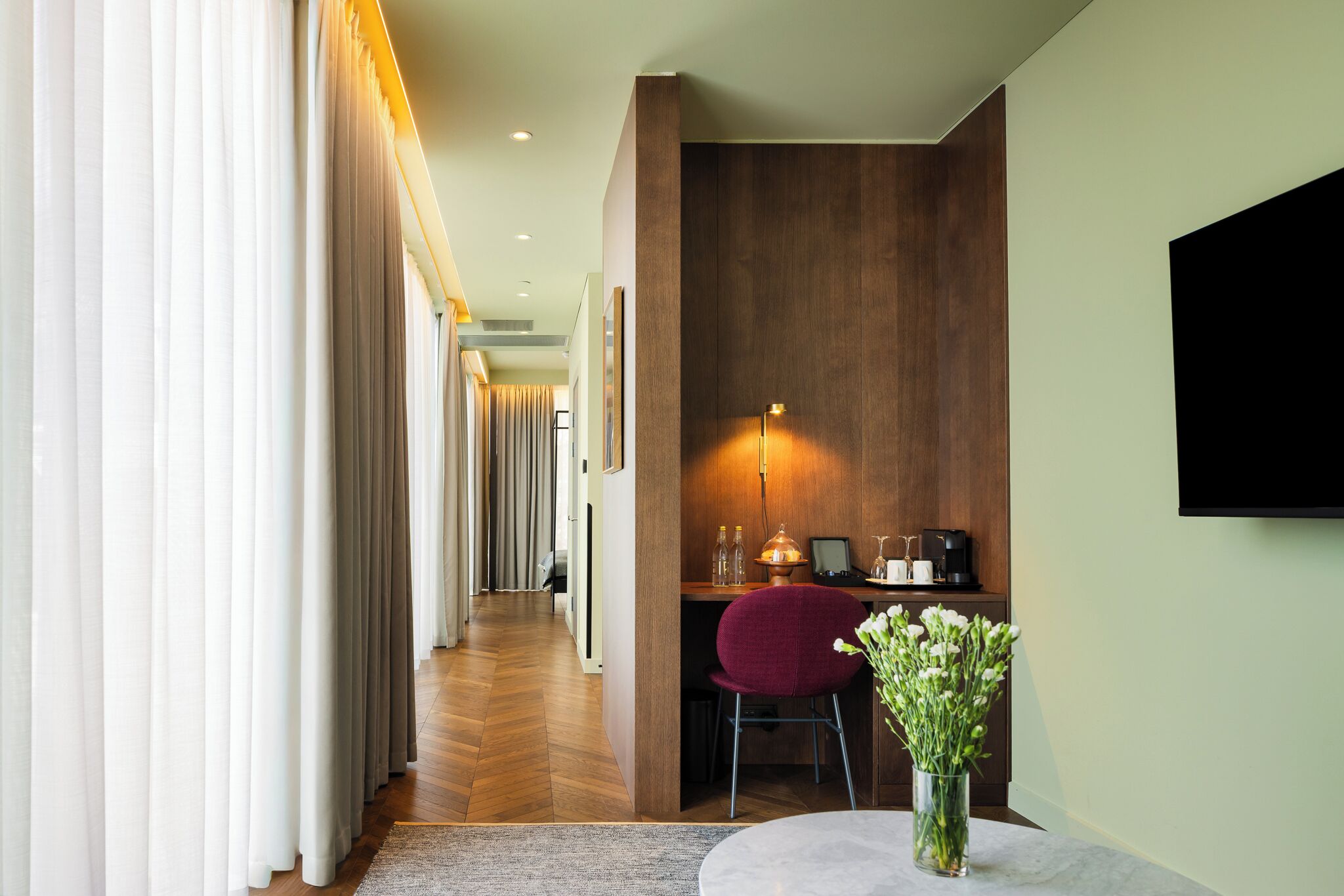 A hotel room with a wooden desk, a chair, a vase with flowers, a flat-screen TV, and a corridor with curtains.