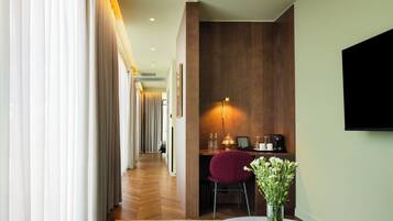A hotel room with a wooden desk, a chair, a vase with flowers, a flat-screen TV, and a corridor with curtains.