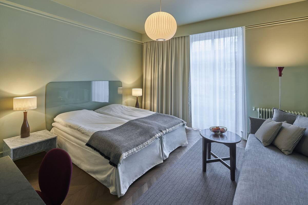 Ein Hotelzimmer mit Bett, Nachttischen, einer Couch, einem kleinen Tisch und einem großen Fenster mit Vorhängen.