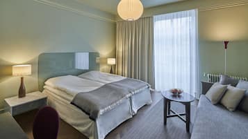 A hotel room with a bed, bedside tables, a sofa, a small table, and a large window with curtains.