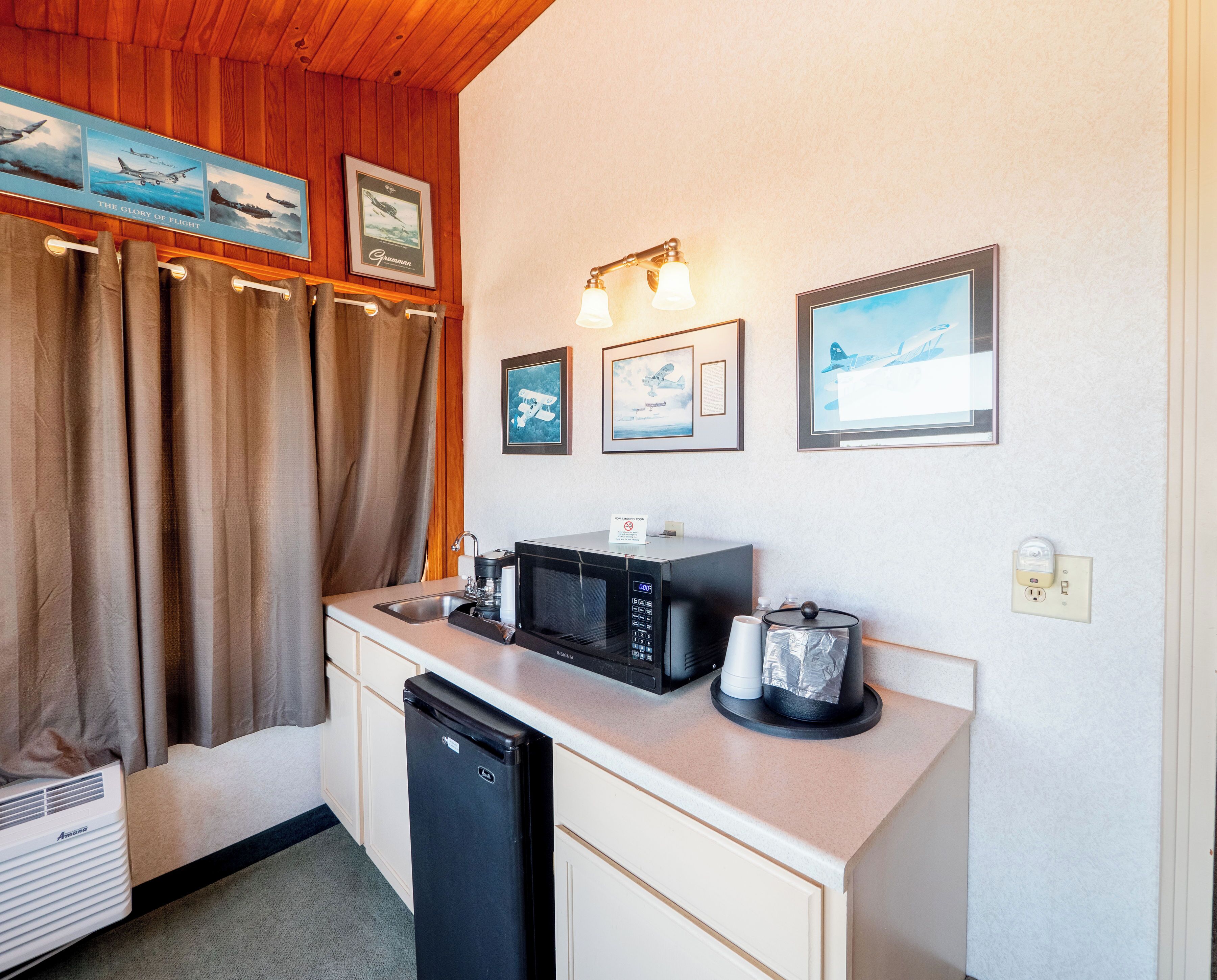 Una kitchenette compacta con microondas, cafetera y un refrigerador pequeño. Hay cuadros enmarcados en la pared, una ventana con cortinas y un techo con paneles de madera.