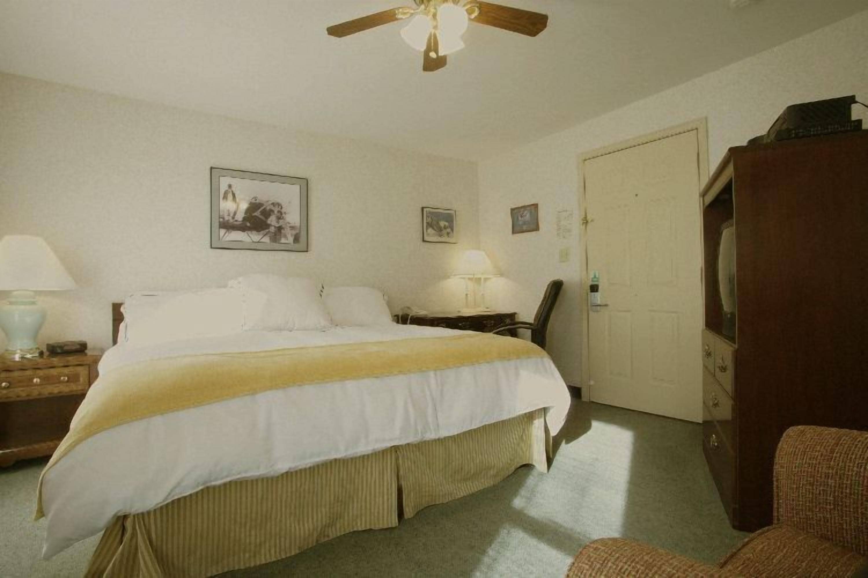 Un dormitorio con una cama, dos mesitas de noche con lámparas, un ventilador de techo, una puerta y un armario de madera.