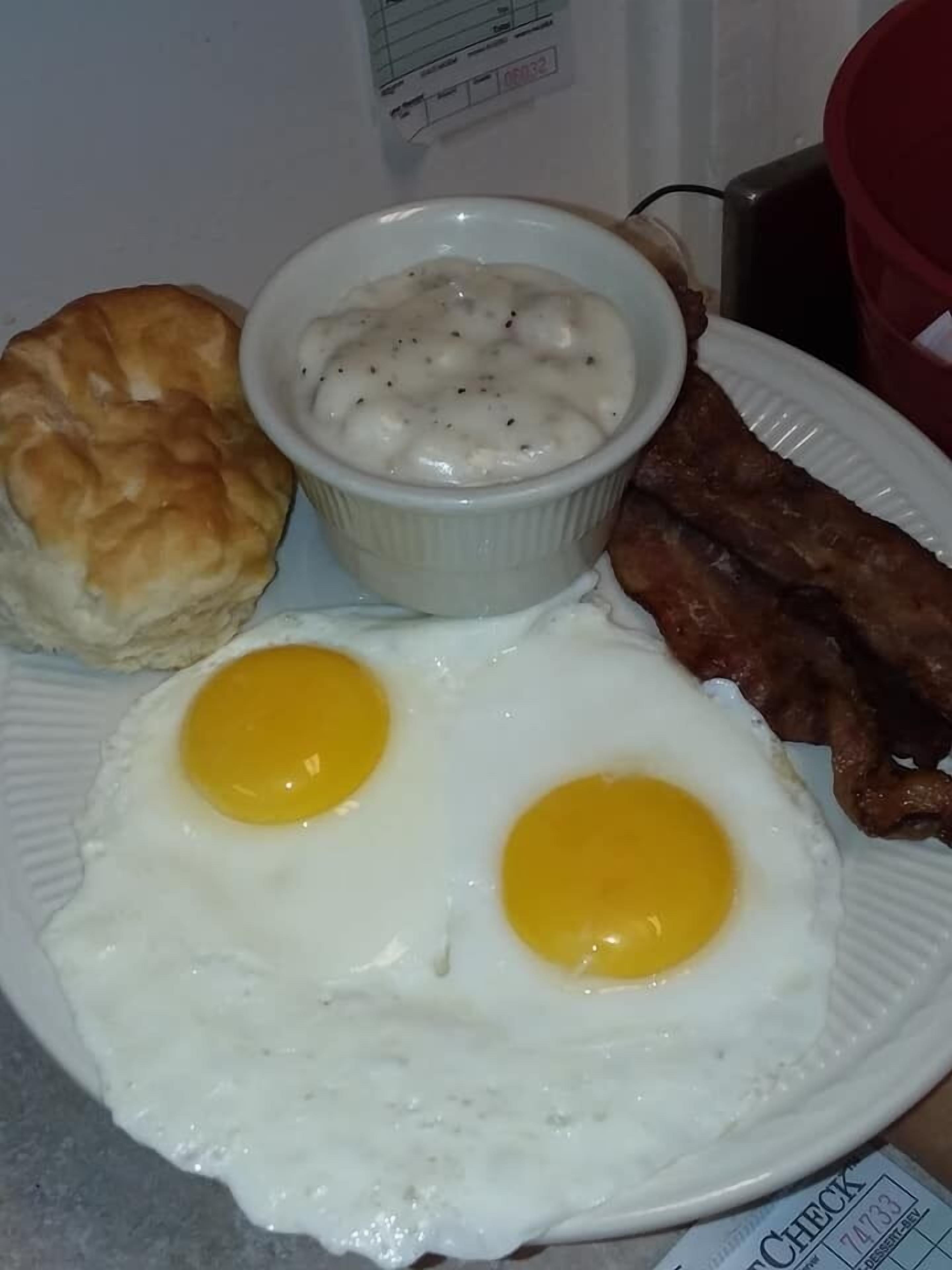 A plate with two fried eggs, a biscuit, a bowl of grits, and bacon.