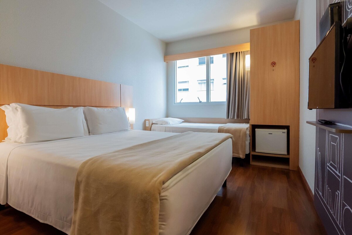 A hotel room with a large bed, a smaller bed, a TV, a wardrobe, and a window with curtains.