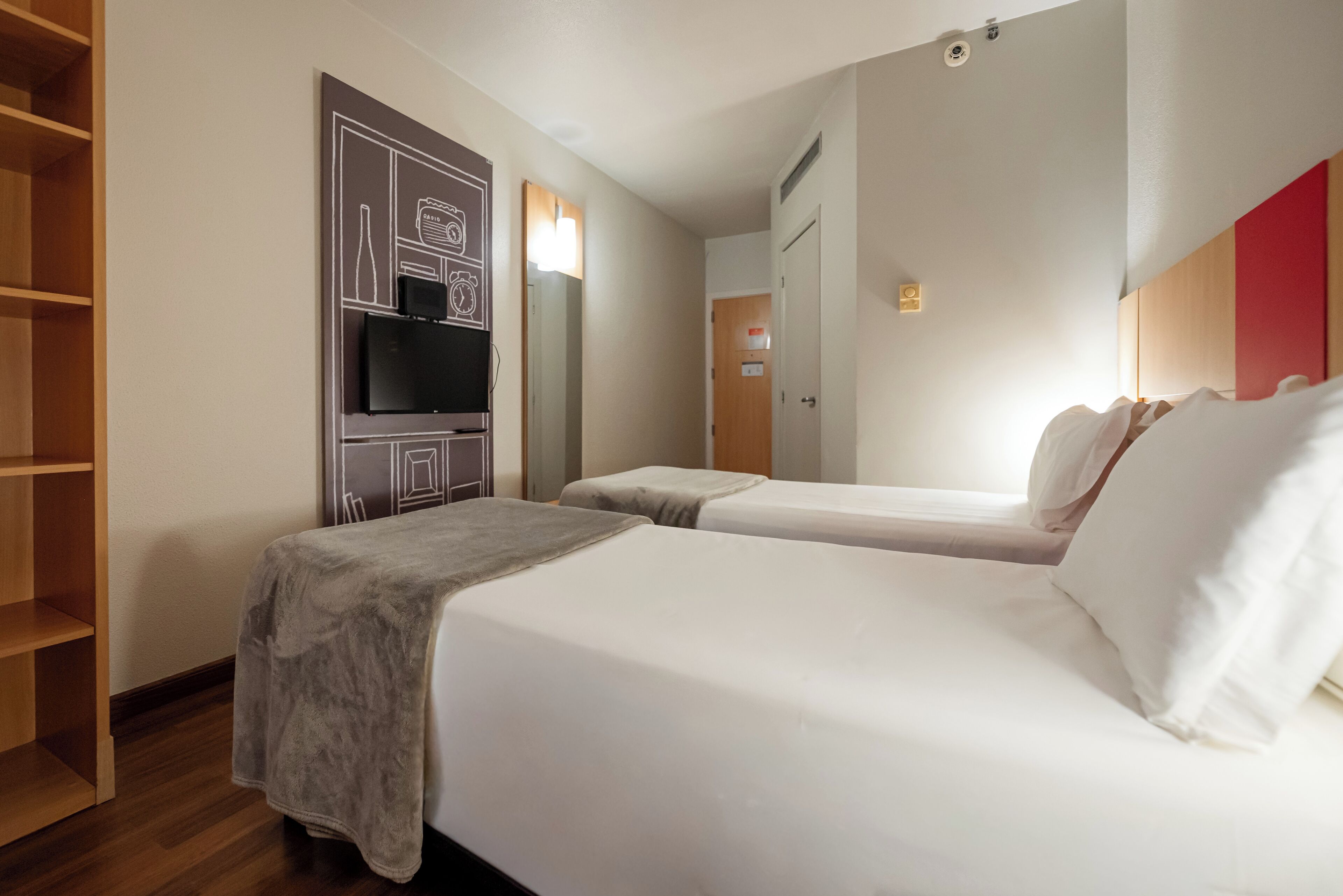 Une chambre d’hôtel avec deux lits, un téléviseur fixé au mur et une étagère en bois.