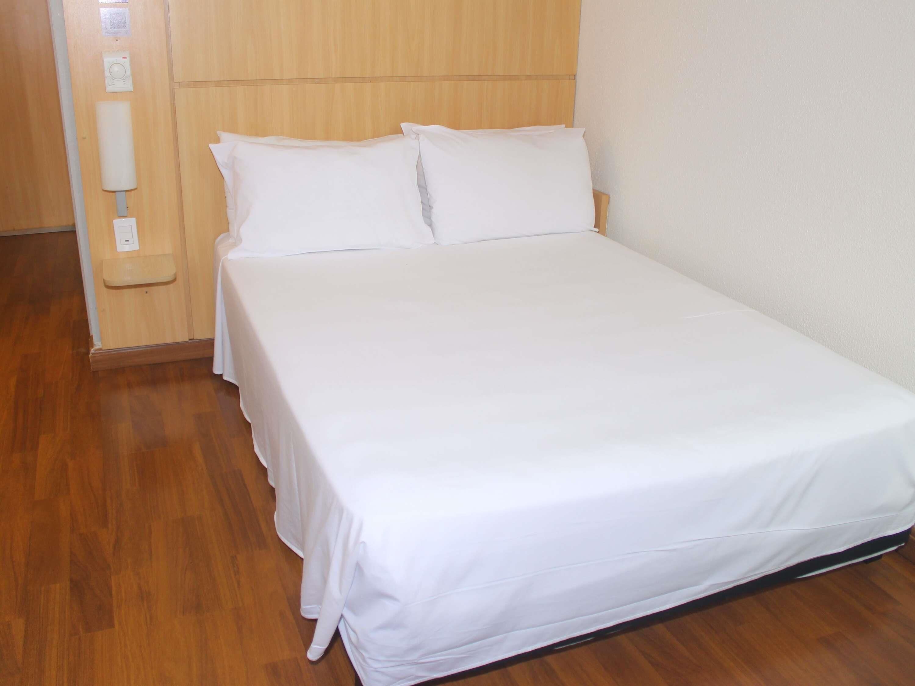 Una cama individual con sábanas blancas en una habitación con suelo de madera y cabecera de madera.
