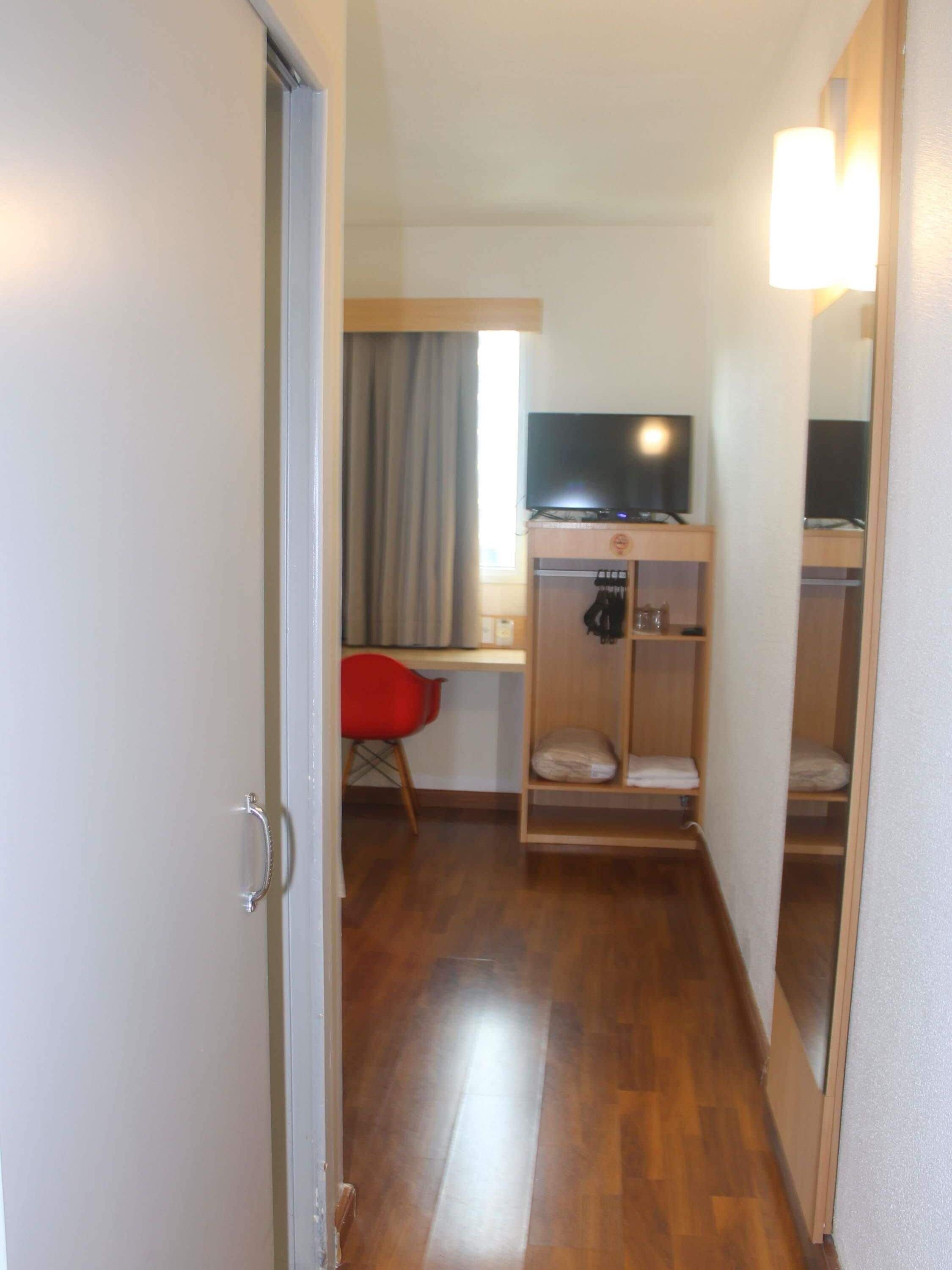 Una habitación de hotel con piso de madera, un escritorio con televisor, una silla roja y un armario.