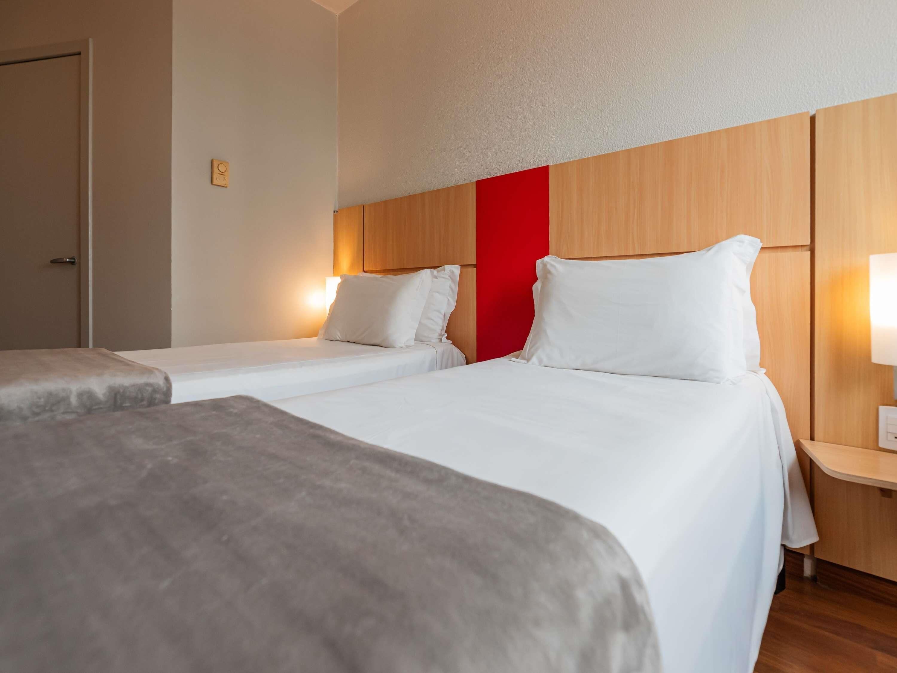 Una camera d'albergo con due letti, una testiera in legno, una parete di colore rosso e un comodino con una lampada.