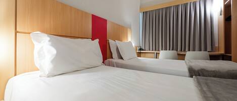 A hotel room with two beds, a wooden headboard, a red accent wall, a desk, and a window with curtains.