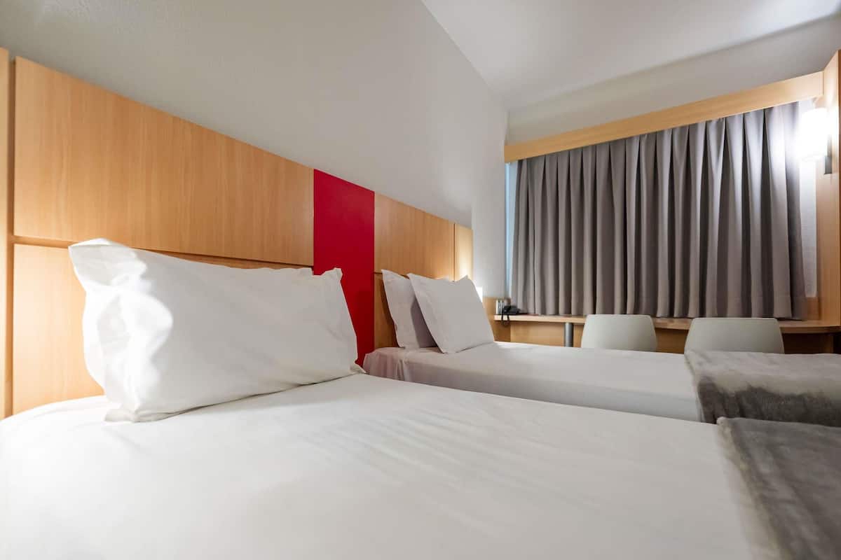 Habitación de hotel con dos camas, cabecero de madera, pared de acento roja, escritorio y ventana con cortinas.