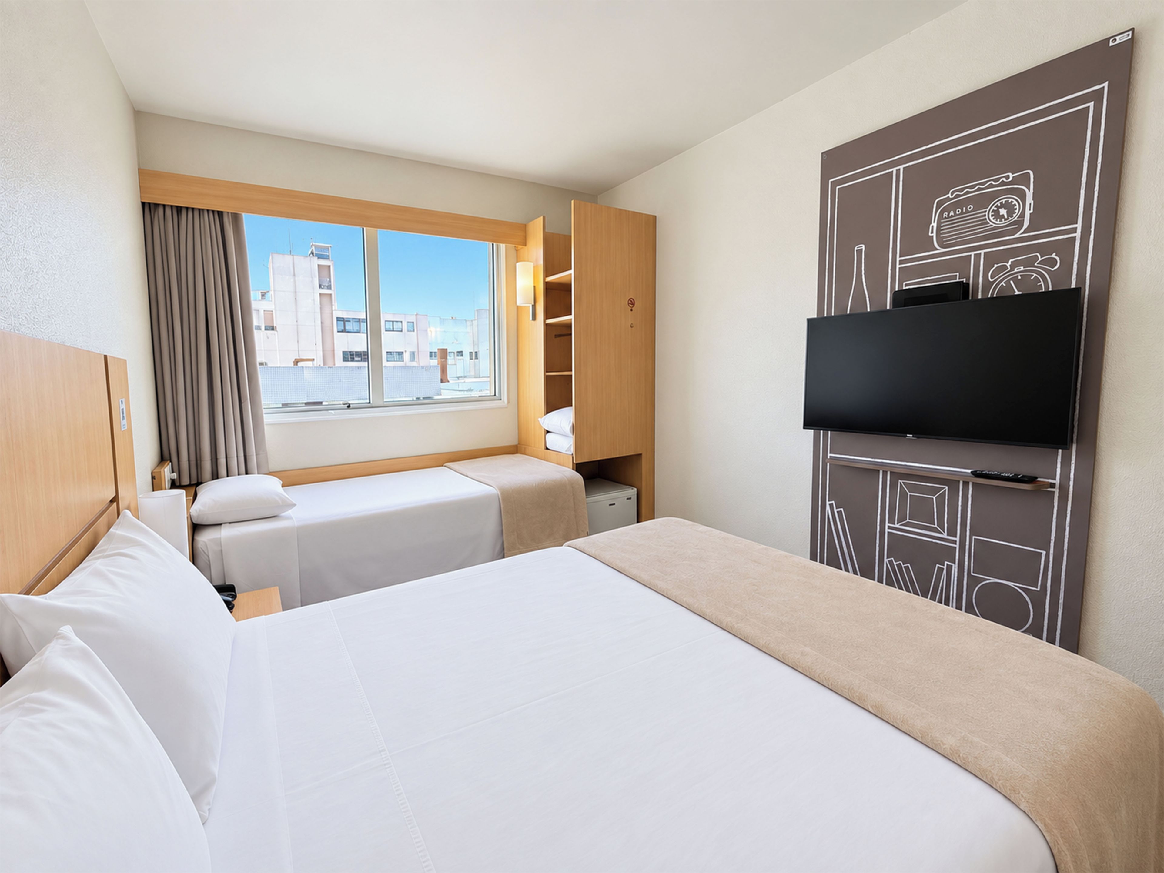 Quarto de hotel com duas camas, televisão, janela com cortinas e vista para edifícios.